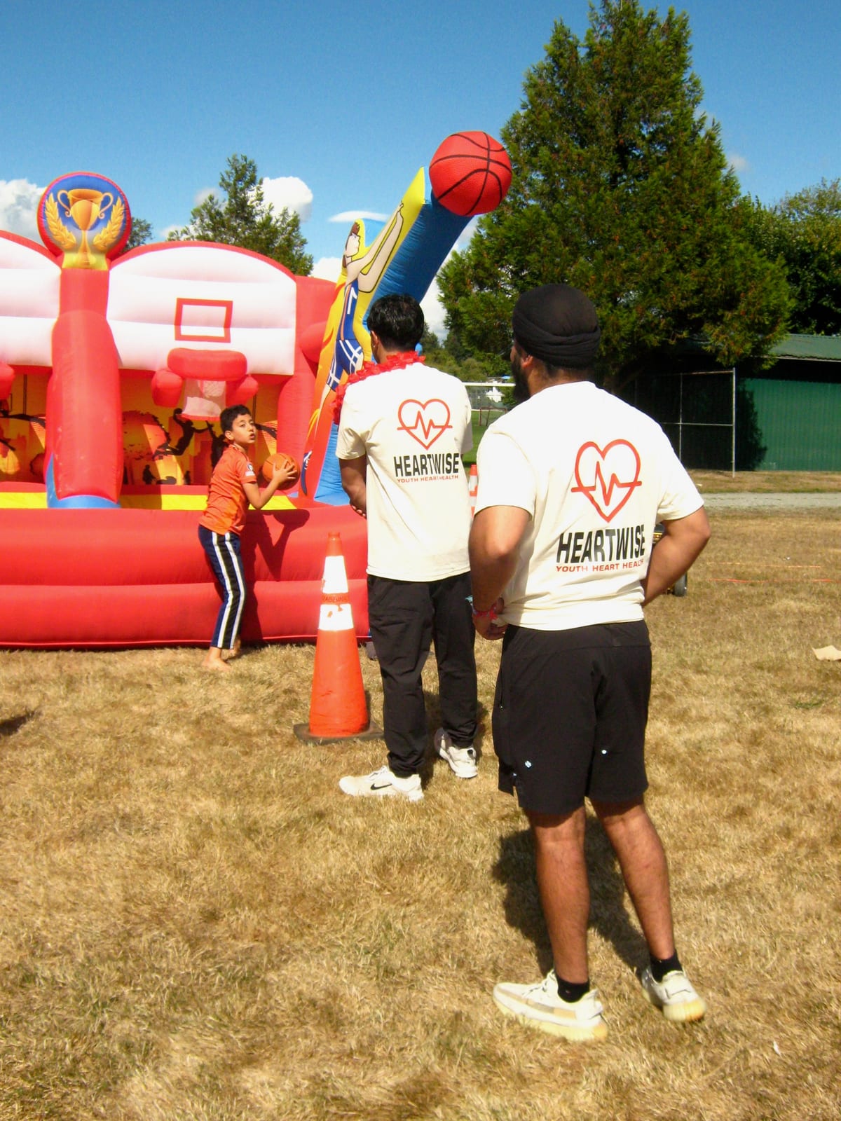 Two volunteers in HeartWise T-shirts watch a small child at the mouth of the red inflatable basketball game.
