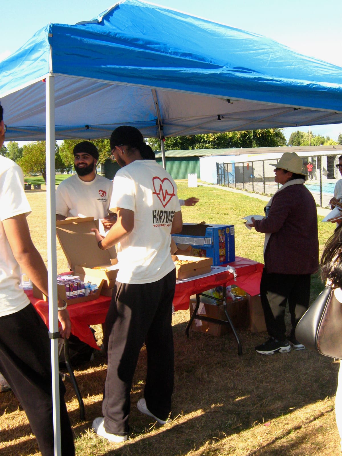 Under a blue pop-up canopy, volunteers unpack boxes at a red-clothed table while a woman in a wide straw hat looks on.