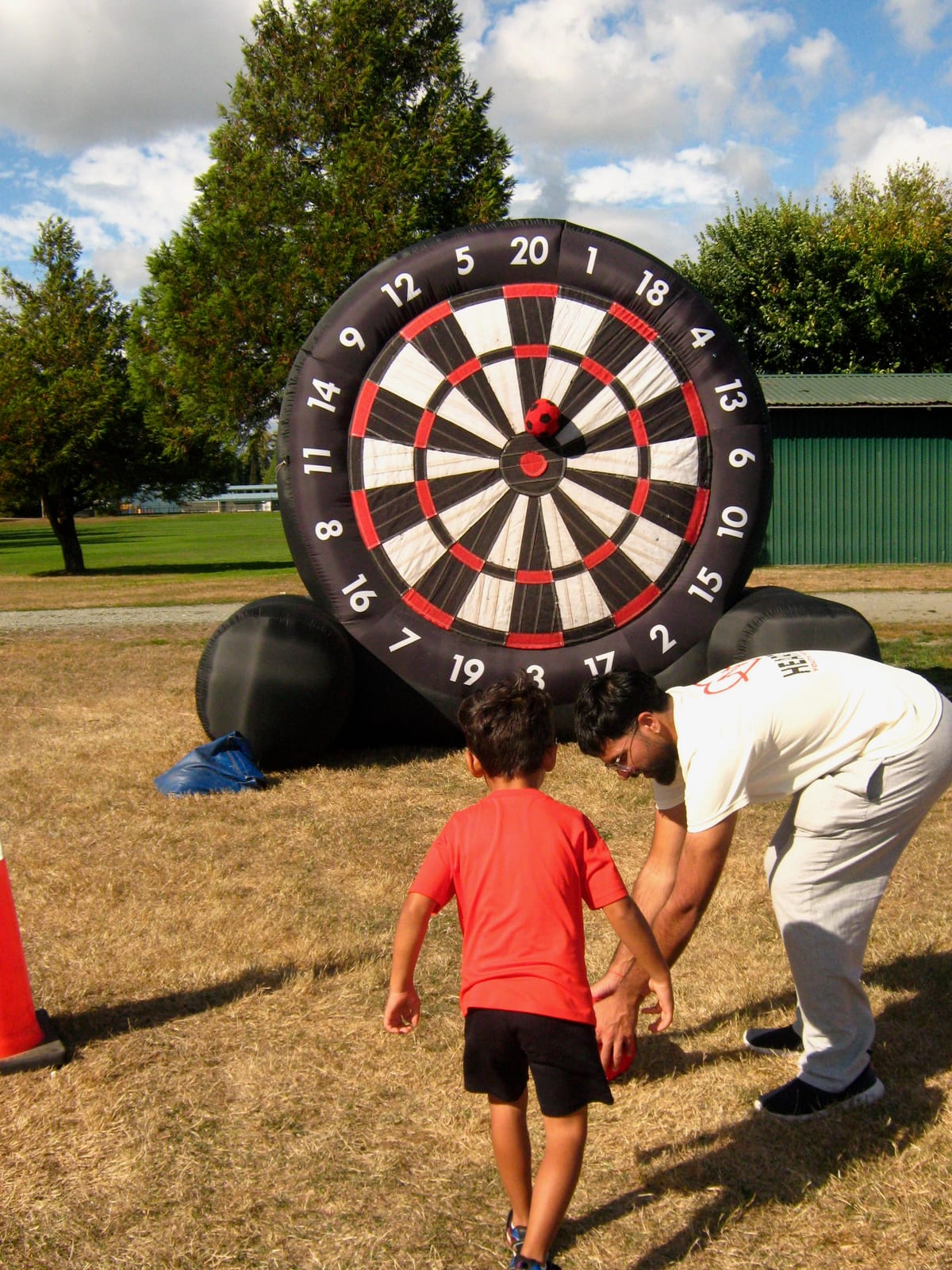 A boy in a red T-shirt faces the giant inflatable dartboard while a volunteer bends to collect the balls.