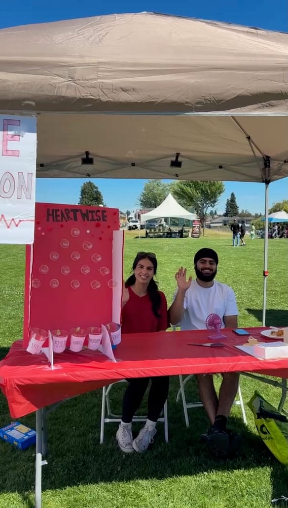 Two volunteers wave from behind the red booth table and the HeartWise peg-board game.