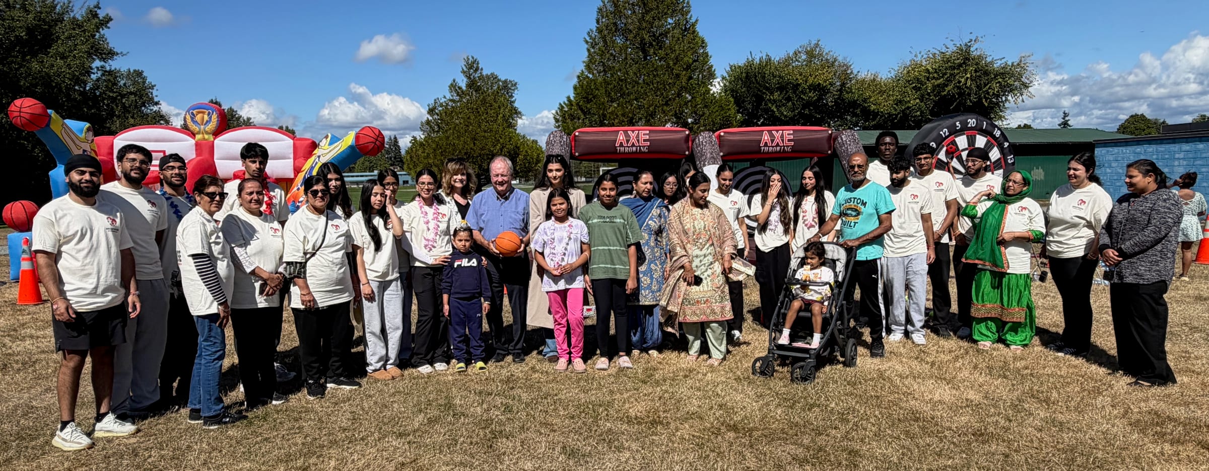 Around forty volunteers, families and guests line up across the field at Unwin Park for a group photo, most in cream HeartWise T-shirts, with the inflatable games behind them under a blue sky.