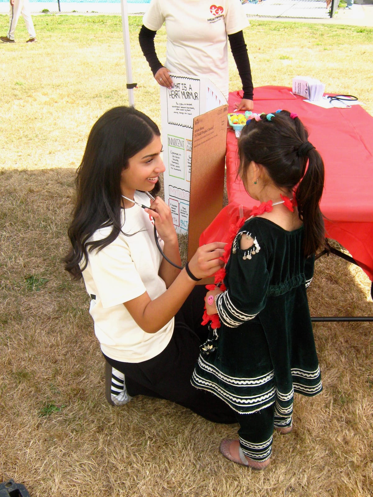 A HeartWise volunteer kneels on the grass and listens to a young child's heart with a stethoscope, beside a hand-made 'What is a heart murmur?' board at Pulse & Play 2026.