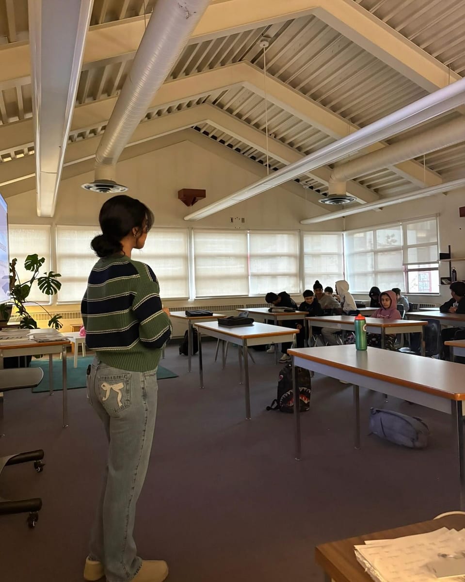 A classroom of students seated at tables, seen from the back, facing a screen at the front.