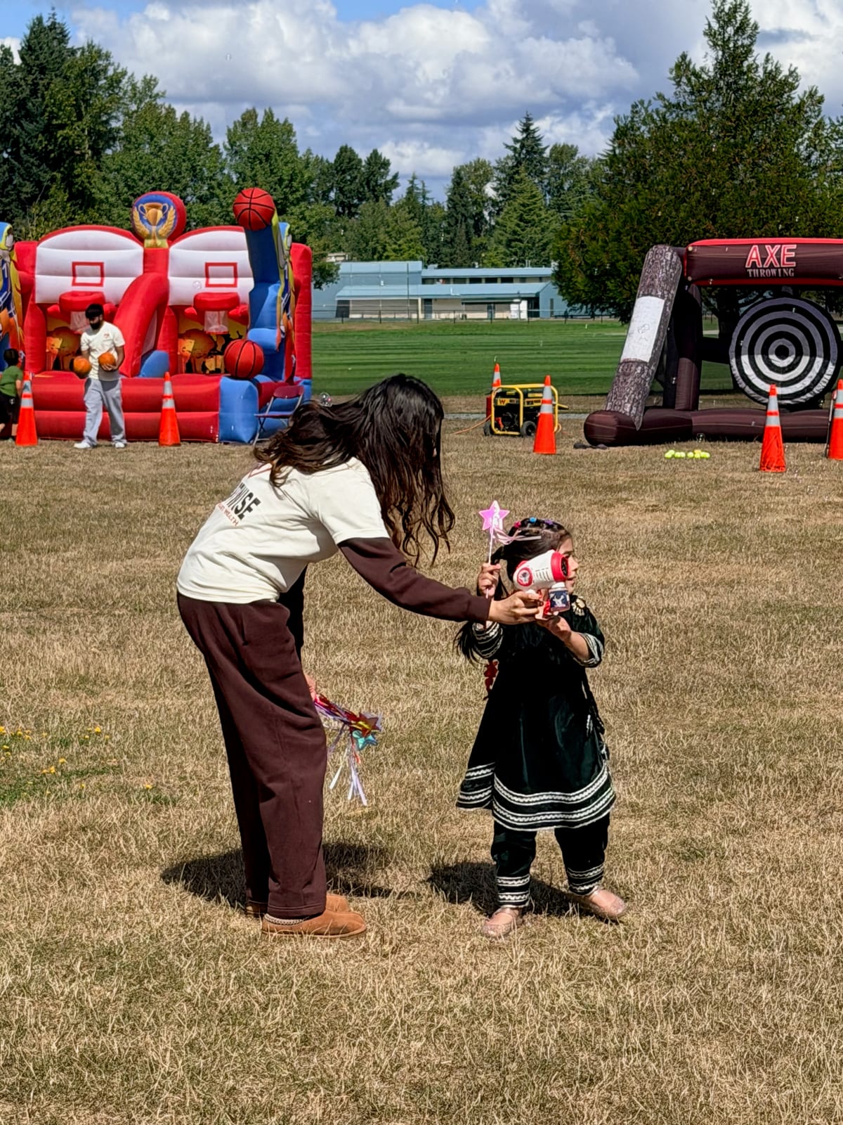 A volunteer in a cream HeartWise T-shirt bends toward a small child holding a pink star wand and a toy megaphone, with the inflatable basketball and axe-throwing games behind them.